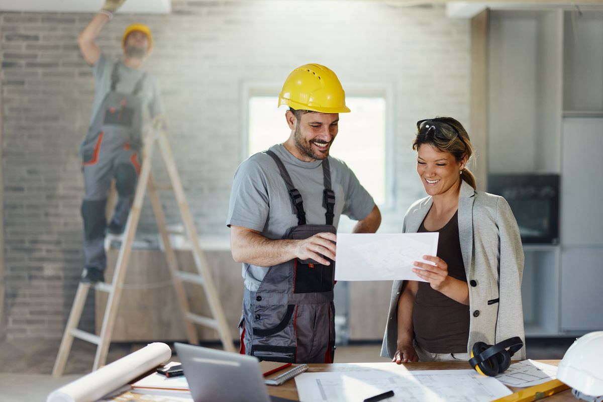 Happy interior designer and manual worker cooperating while analyzing housing plans during home renovation process.