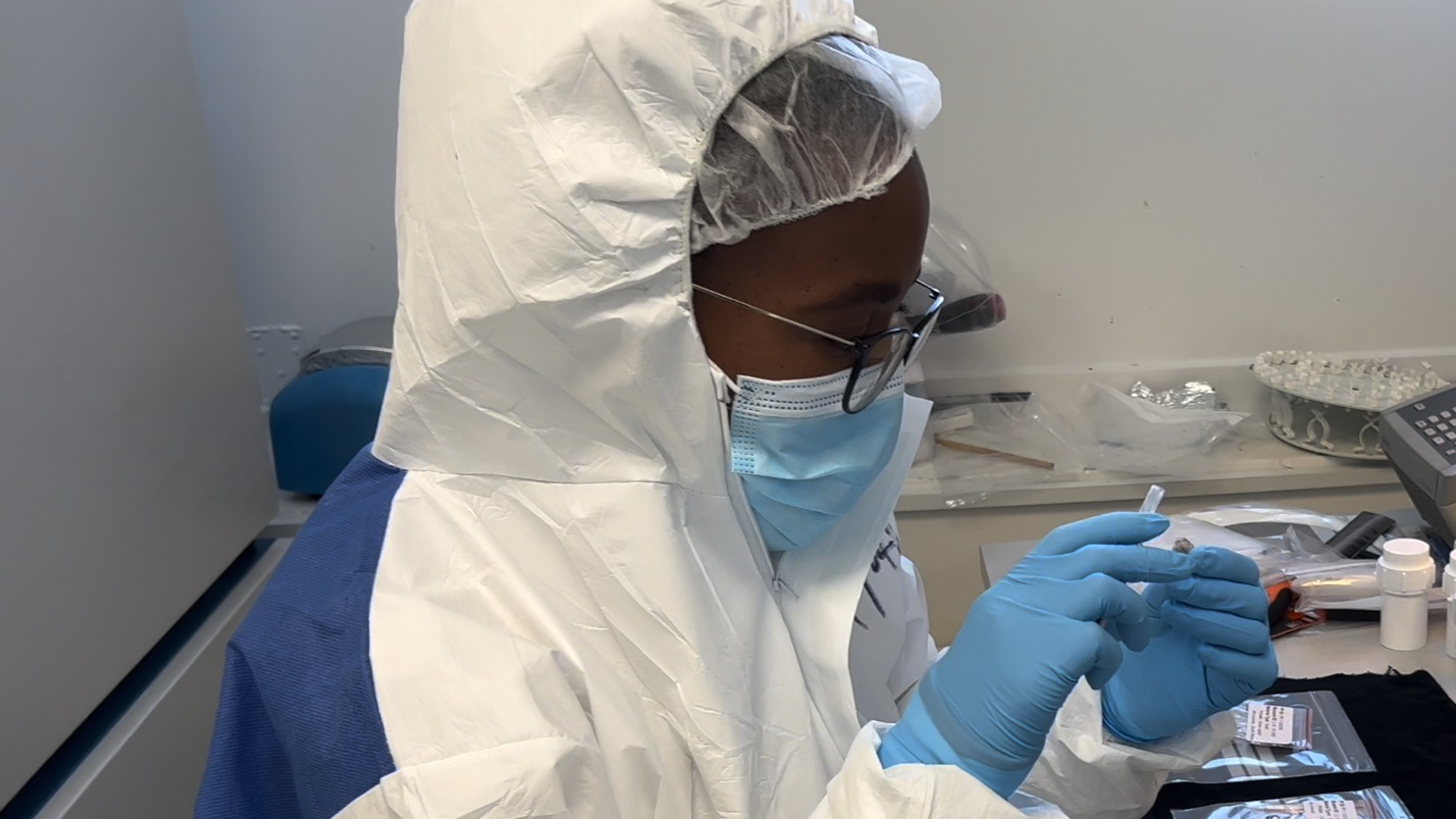a scientist with dark skin and glasses wears white scrubs and blue gloves to test something