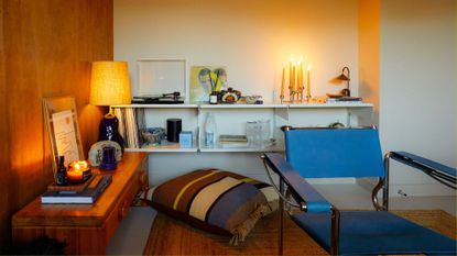 A white living room with a wood accent wall, floating white shelves, a wood sideboard, a table lamp, a turntable, a cluster of white taper candles, a striped floor cushion, and a blue chair