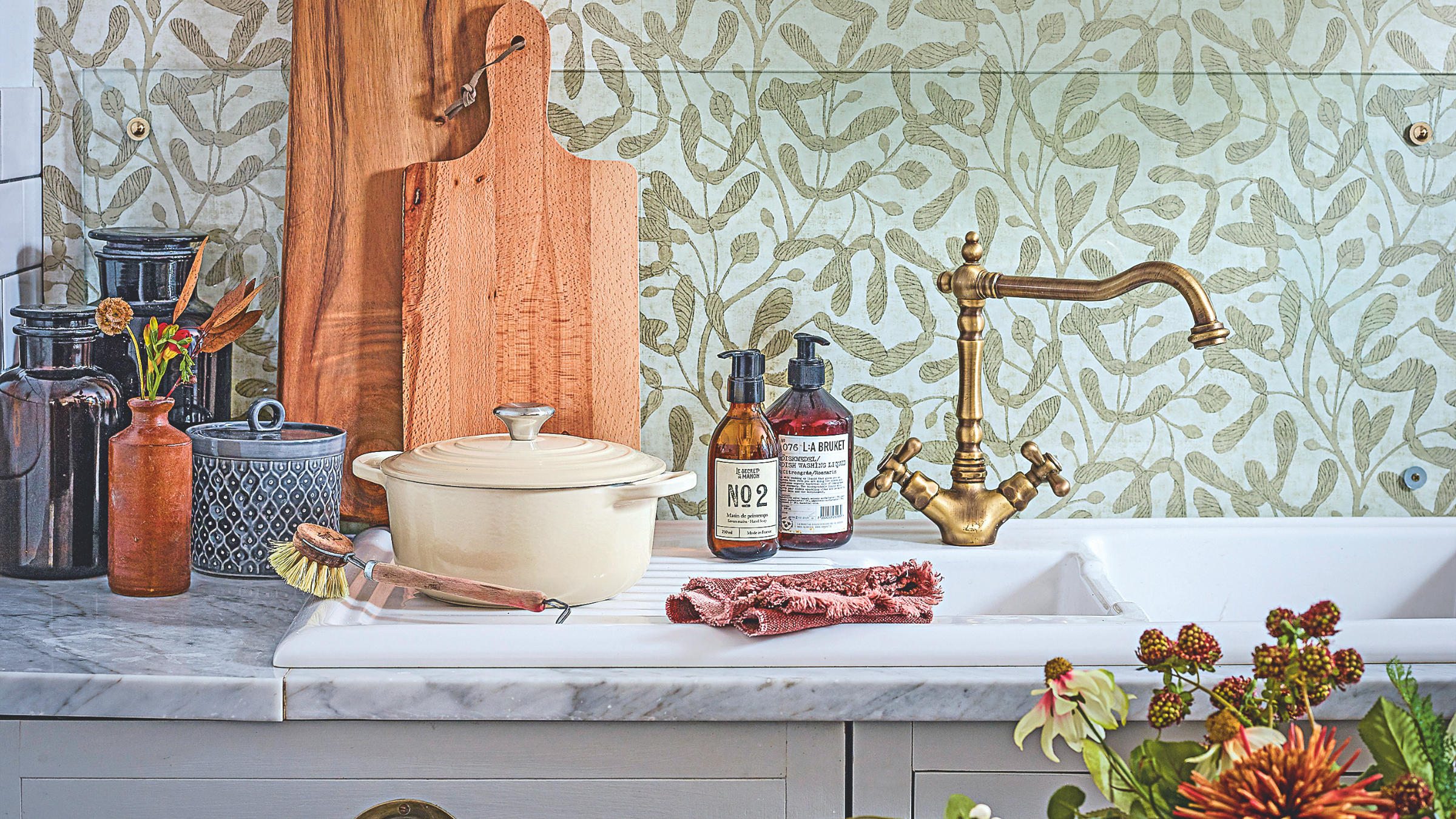 Kitchen sink and draining board in front of mistletoe wallpaper on a marble worktop with a stool decorated with flowers.