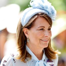 Carole Middleton wearing a blue dress with a tie neck and a blue headband with a large flower smiling and looking to the right 