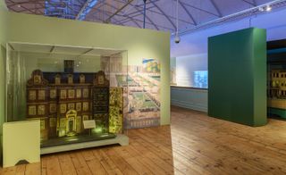 View of a multi-level, brown doll house in a glass display case at the V&A Museum of Childhood. The space features wood flooring and green walls with wall art