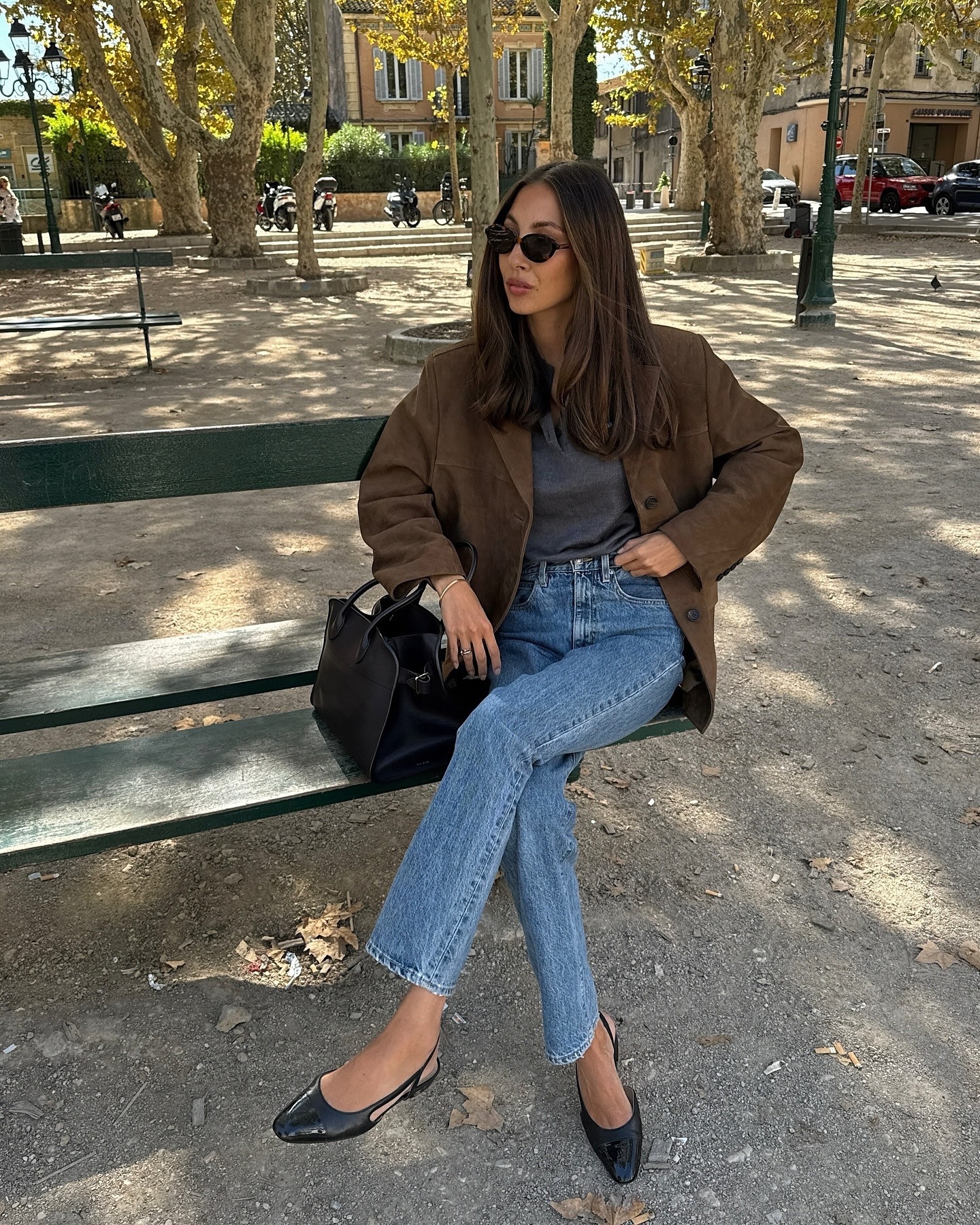 style influencer Felicia Akerstrom sits on a bench in a Paris park wearing black sunglasses, a brown suede jacket, gray top, high-waisted straight-leg jeans, a black top handle bag, and black slingback flats