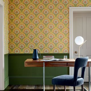 a desk and chair beside a wall painted forest green below the dado rail and covered in a bold yellow and green print wallpaper above