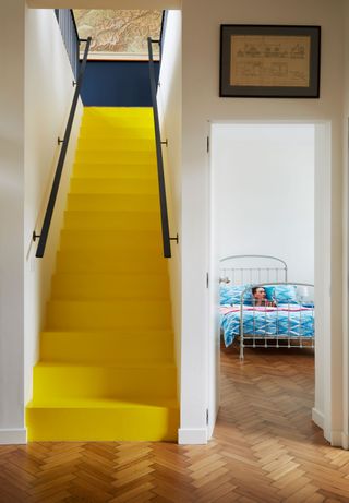 A bright yellow painted staircase with black handrails in white hallway with Herringbone-style wooden flooring with white bed frame with blue duvet in background