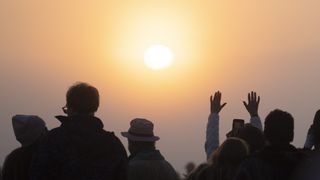 view of the sun shining through hazy cloud with people in the foreground looking towards the sun, one person has both of their hands raised to the sky.