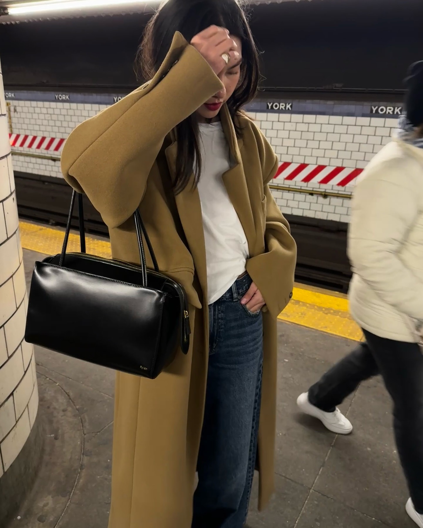 NYC based style influencer Mimi Nguyen poses on a Subway platform wearing an oversized mustard green coat, a white t-shirt, jeans, and a black bag