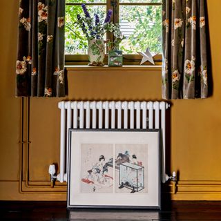 A window with floral curtains and a radiator, surrounded by a yellow wall