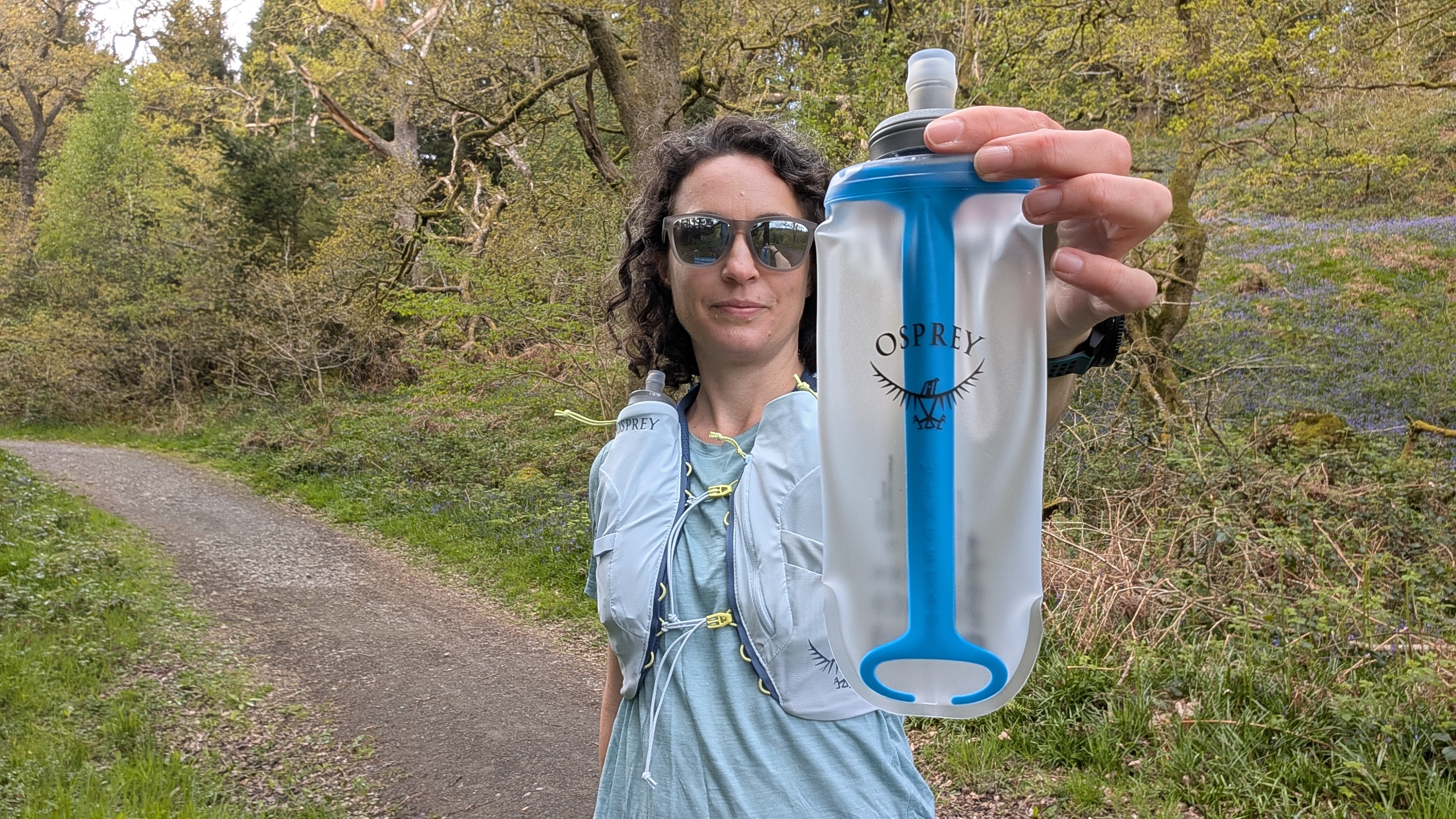 A woman trail runner wearing a light blue T-shirt and holding up the water bottle from her Osprey Dyna running vest