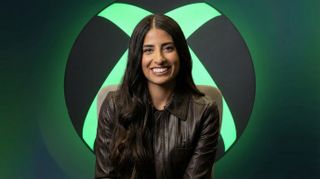 A composited image of Asha Sharma seated in front of a large illuminated Xbox logo on a clean gradient background.