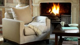 Beige armchair pulled up beside an open fire to create a cozy nook as an idea for how to make a home cozy during the winter