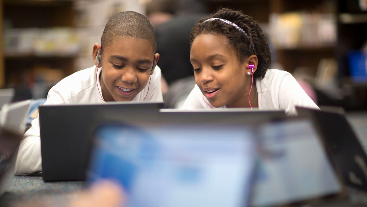 Two students with headphones in leaning in to look at laptop screens in an IT lesson, with more laptops closer to the camera but out of focus.