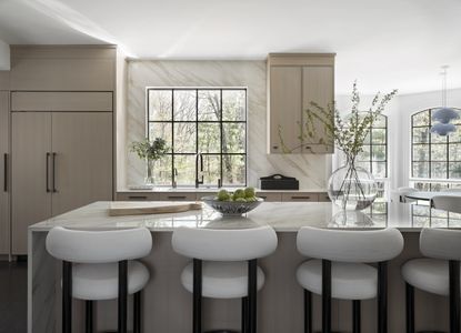 A neutral kitchen with a marble island, wing-backed bar stools and large windows with black trim