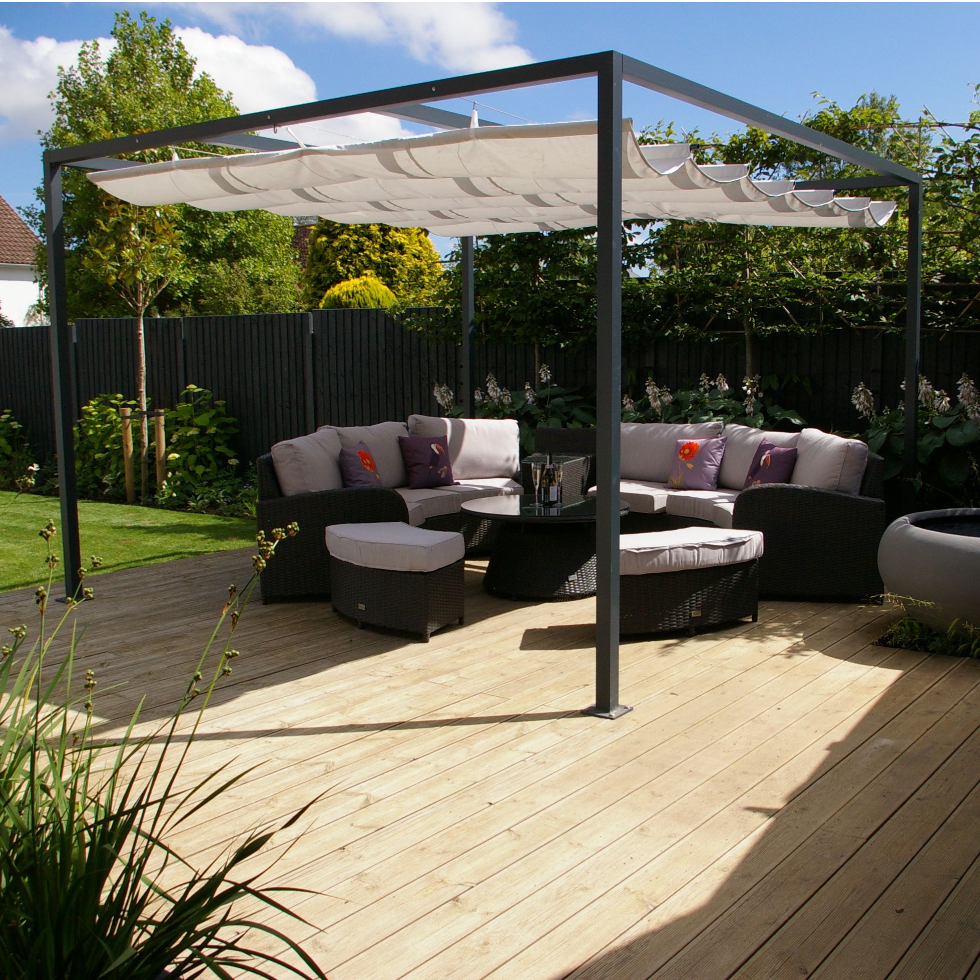 A black metal pergola with a retractable canopy above a black garden furniture set with white cushions