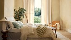 White and green linens on a bed against cream walls and curtains looking onto a garden view. 