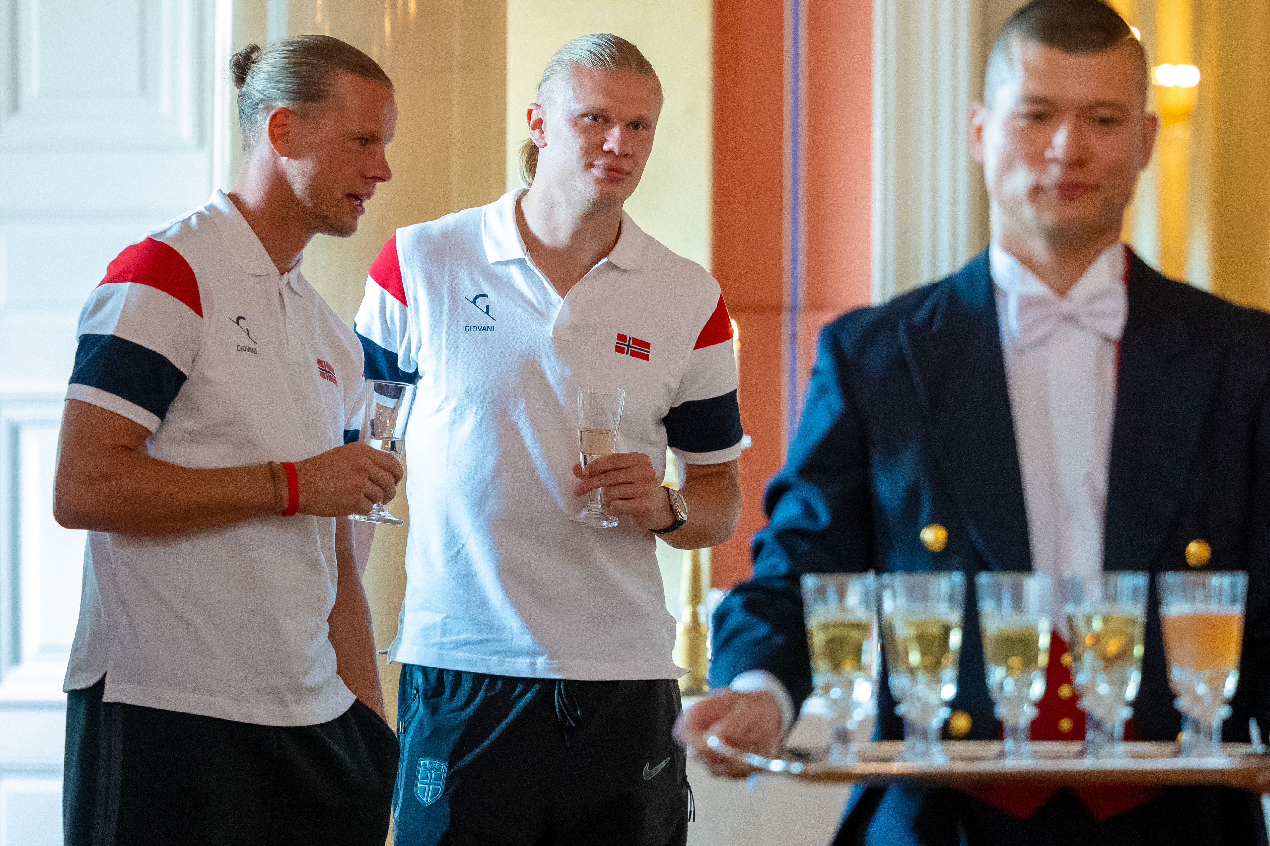Erling Haaland standing behind a man carrying champagne on a tray