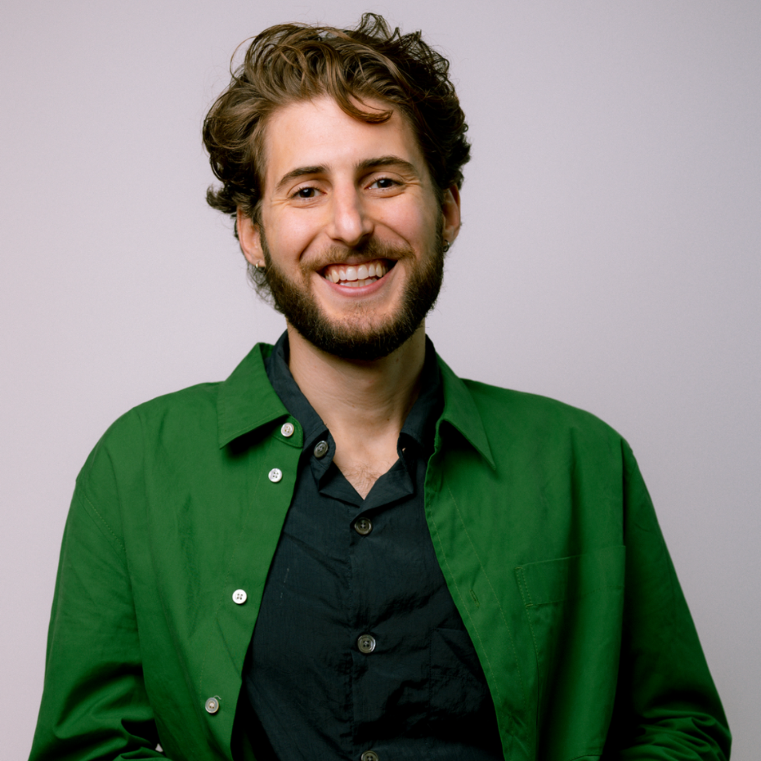 A smiling man with brown hair and a beard in a green unbuttoned shirt and a dark green shirt