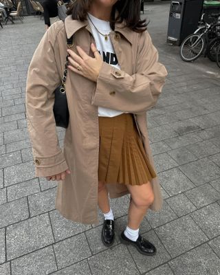 Francesca wears a beige trench coat, pleated mini skirt and loafers