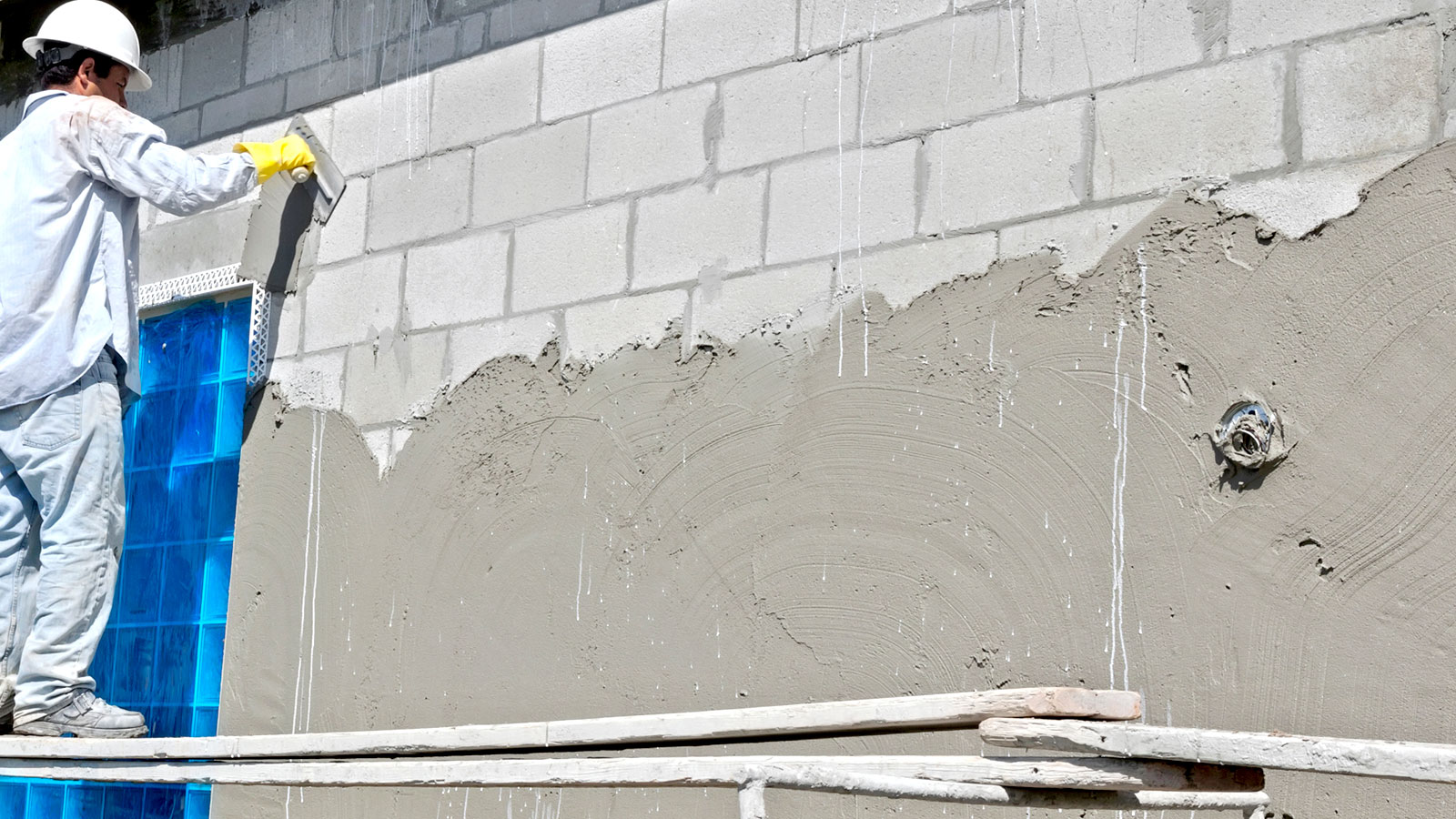 Plasterer on raised platform applying grey stucco plaster to exterior block wall