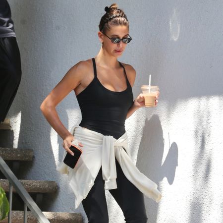 Hailey Bieber wears flared leggings with black high-vamp flats and a black tank top with a white jumper tied around her waist.