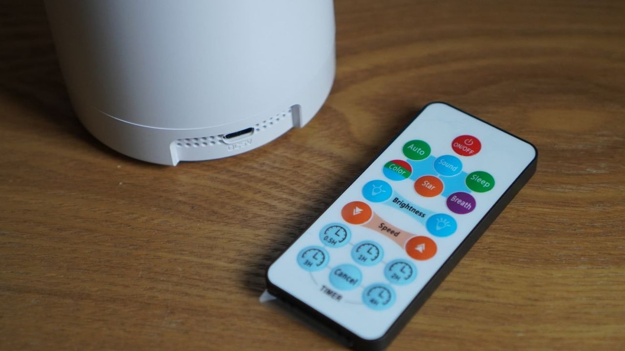 A white spherical projector is next to a multicolor remote control, both on a brown wooden surface.