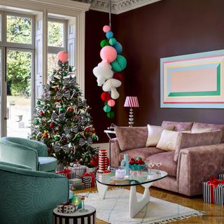 Living room with Christmas tree, striped bows and pastel ribbons, paper decorations hanging down, pink sofa and aqua armchairs, glass coffee table, brown walls, artwork