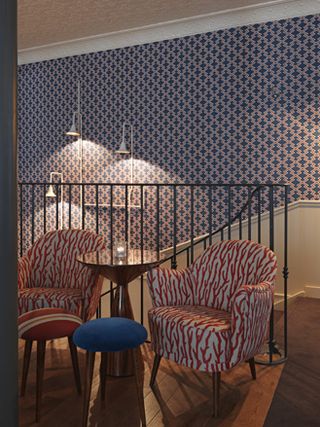 The Fish Club, Paris, France. A sitting area with a round table and patterned chairs above a staircase with patterned wallpaper and wall lights.