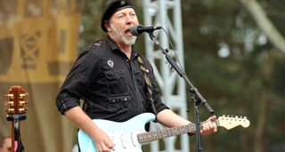 Richard Thompson performs onstage during a daytime festival appearance with a Daphne Blue Fender Stratocaster – he wears a black shirt and black hat.