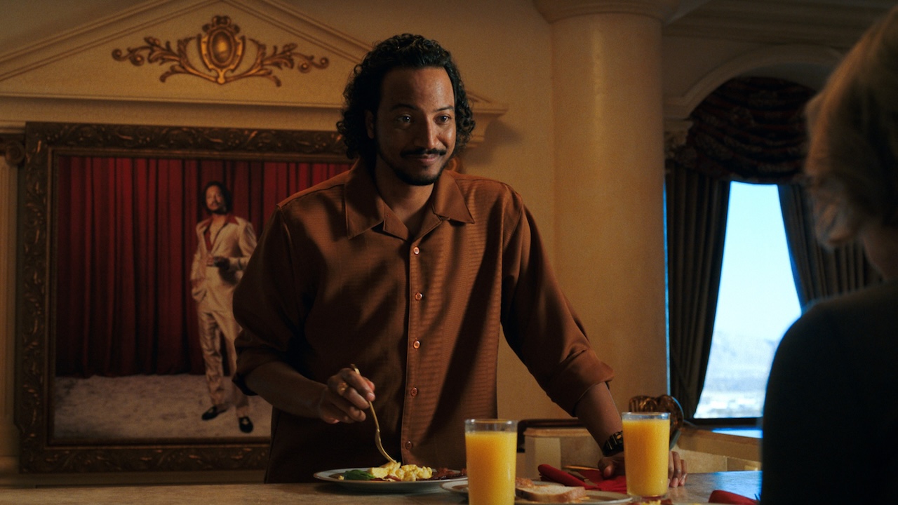 Koumba serving Carol breakfast in front of a portrait of himself in Pluribus Episode 6