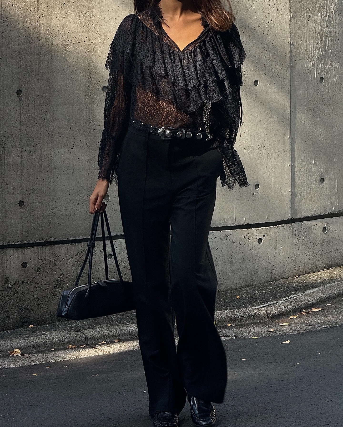 An image of an influencer walking in Japan wearing a black lace long-sleeved peasant blouse, black trousers and black loafers carrying a black baguette bag and wearing a studded belt.