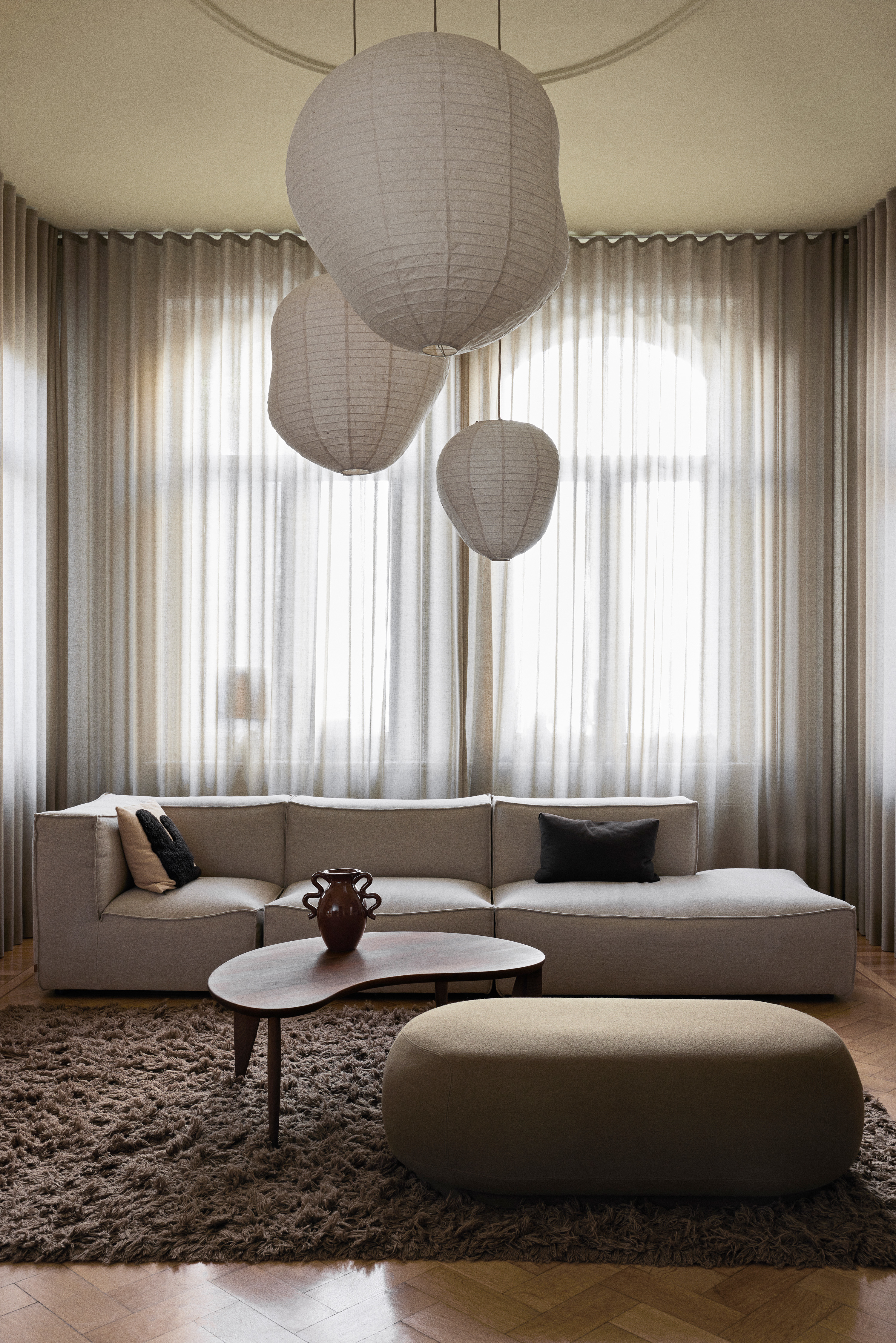 A light contemporary lounge with sheer curtains, a large sofa, coffee table and oversized pouffe