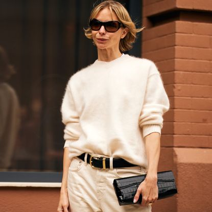 Fashion week spring 2026 attendee wearing sunglasses, white v-neck sweater, black belt, white pants, black clutch