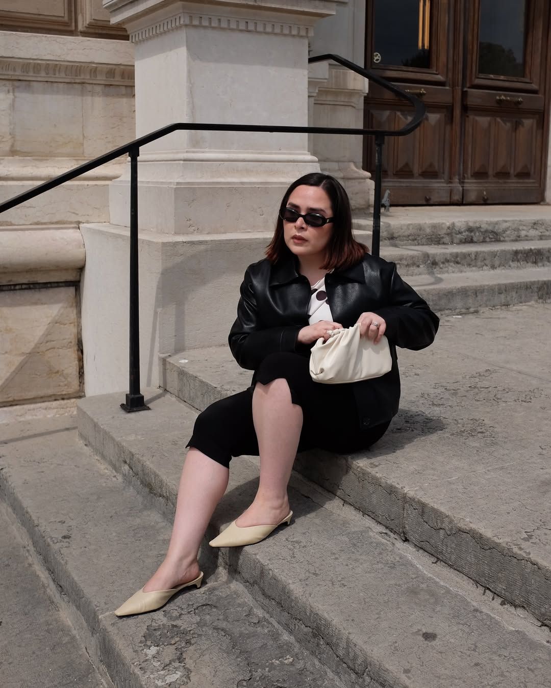 @indiraci wears pale yellow mules with a polka dot top, black capri pants, black leather bomber jacket and white clutch