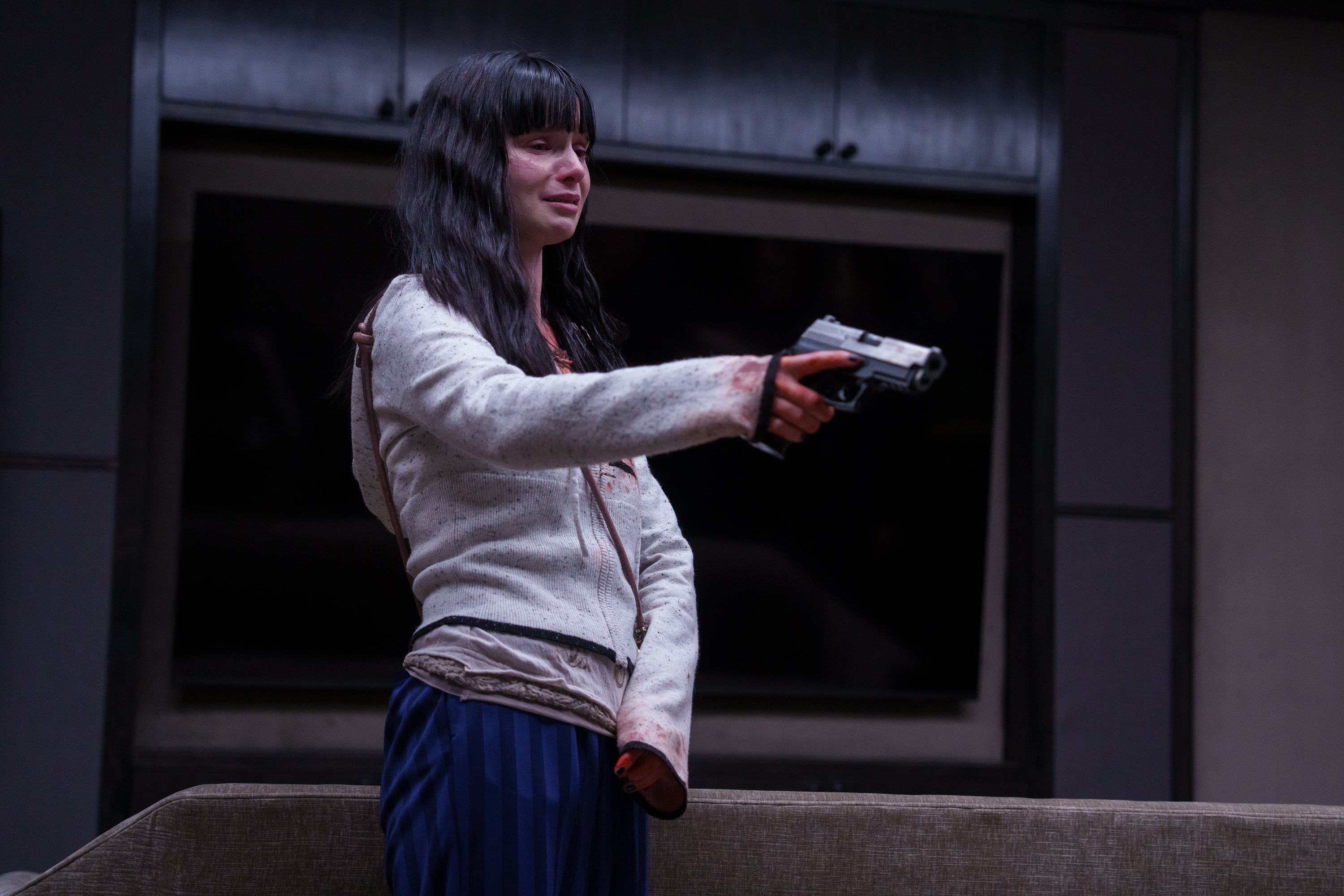A woman in a bloody hoodie (Lola Petticrew as Catherine) points a gun while standing in a home movie theater, in Hulu's 'Furious.'