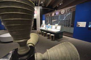 A row of replica Mission Control consoles invite visitors to the Saint Louis Science Center to try their hand directing a (simulated) Apollo moon landing as part of the Smithsonian's "Destination Moon: The Apollo 11 Mission" exhibition. In the foreground are the thrusters from a full-size mockup of the Apollo lunar module ascent stage.