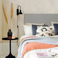 Limewashed walls in bedroom, grey headboard, bed furnished with array of soft furnishings and throws, black bedside table with lamp