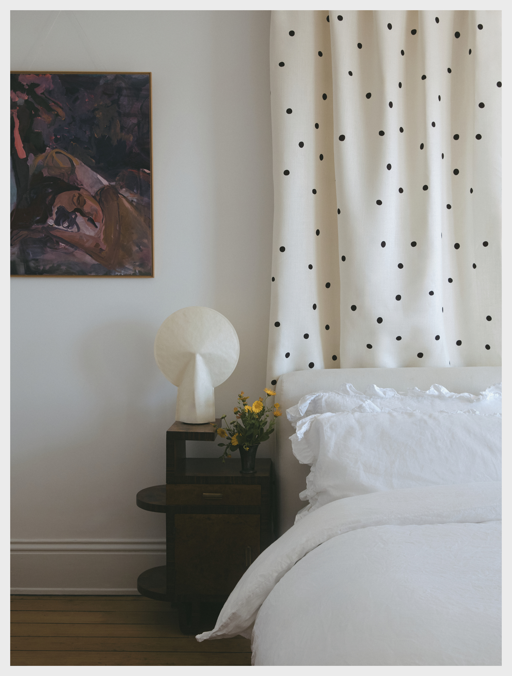 Image of a white bedroom with a white bed and a black-and-white polka dot curtain behind the bed. There is a wooden beside table with a white lamp on it.