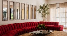 A living room with a modular, red velvet sofa, framed mirrors sitting next to each other on a wall, see-through curtains, and a large beige rug.