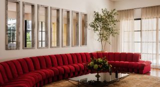 A living room with a modular, red velvet sofa, framed mirrors sitting next to each other on a wall, see-through curtains, and a large beige rug.