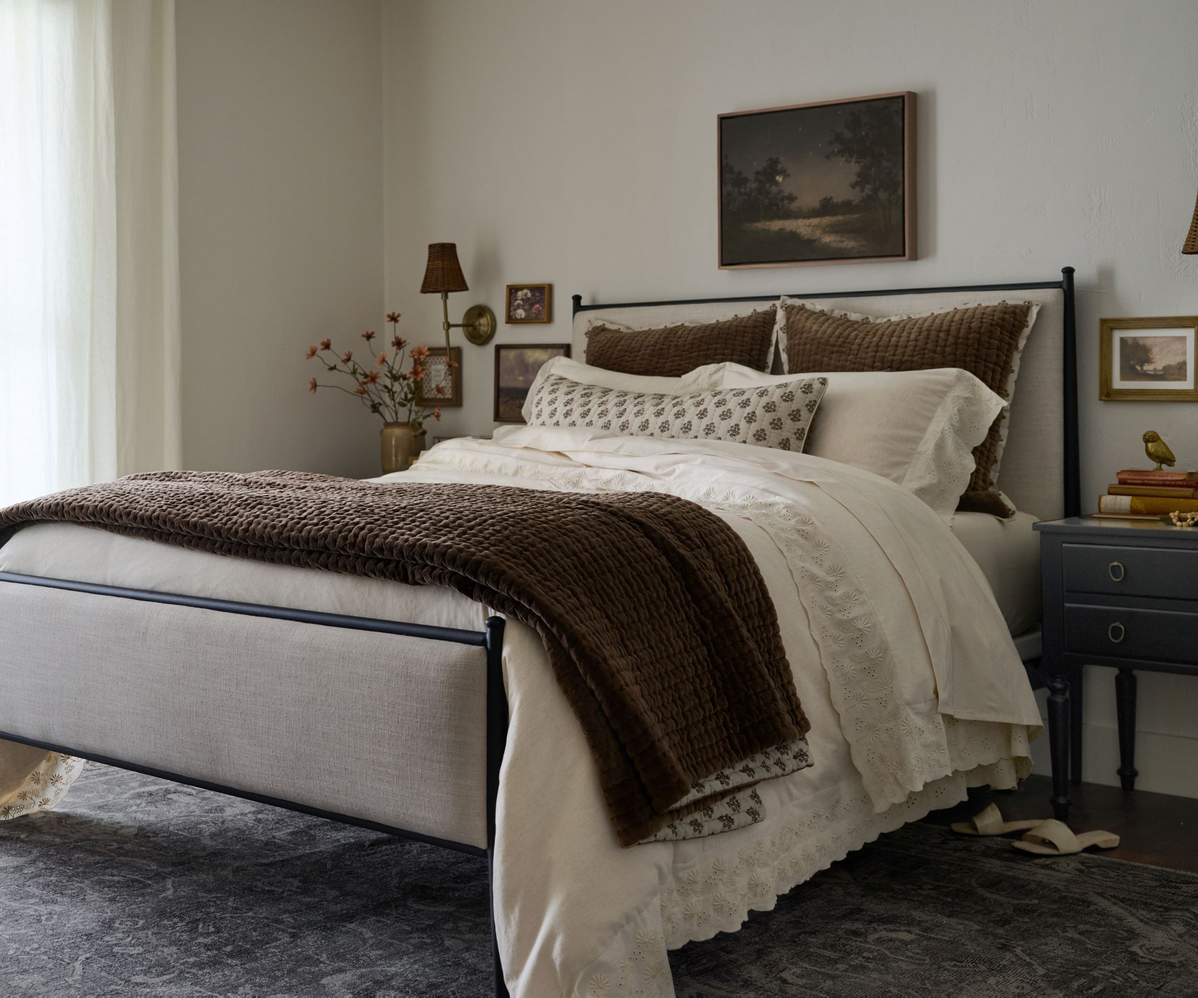 Bedroom with iron bed frame, neutral bedding with brown throw, vintage landscape on wall, side table with books and sconce