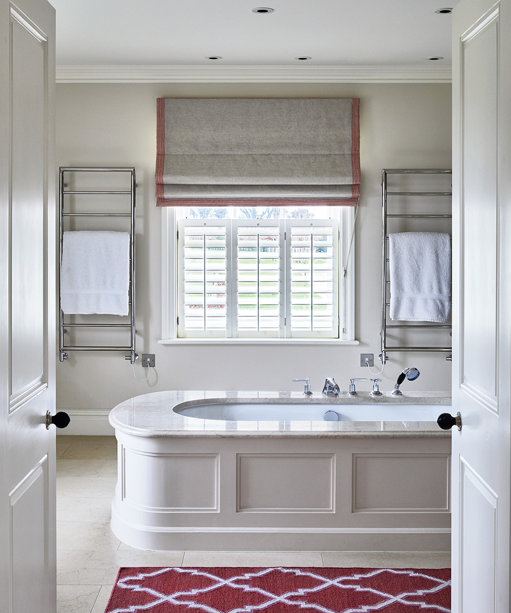 A neutral bathroom with a bath in the centre next to a red and white patterned rug