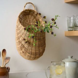 A wicker basket from Magnolia. It's deep and has dried flowers hanging out of it. It's hanging off a white wall with shelves alongside it and a table in front