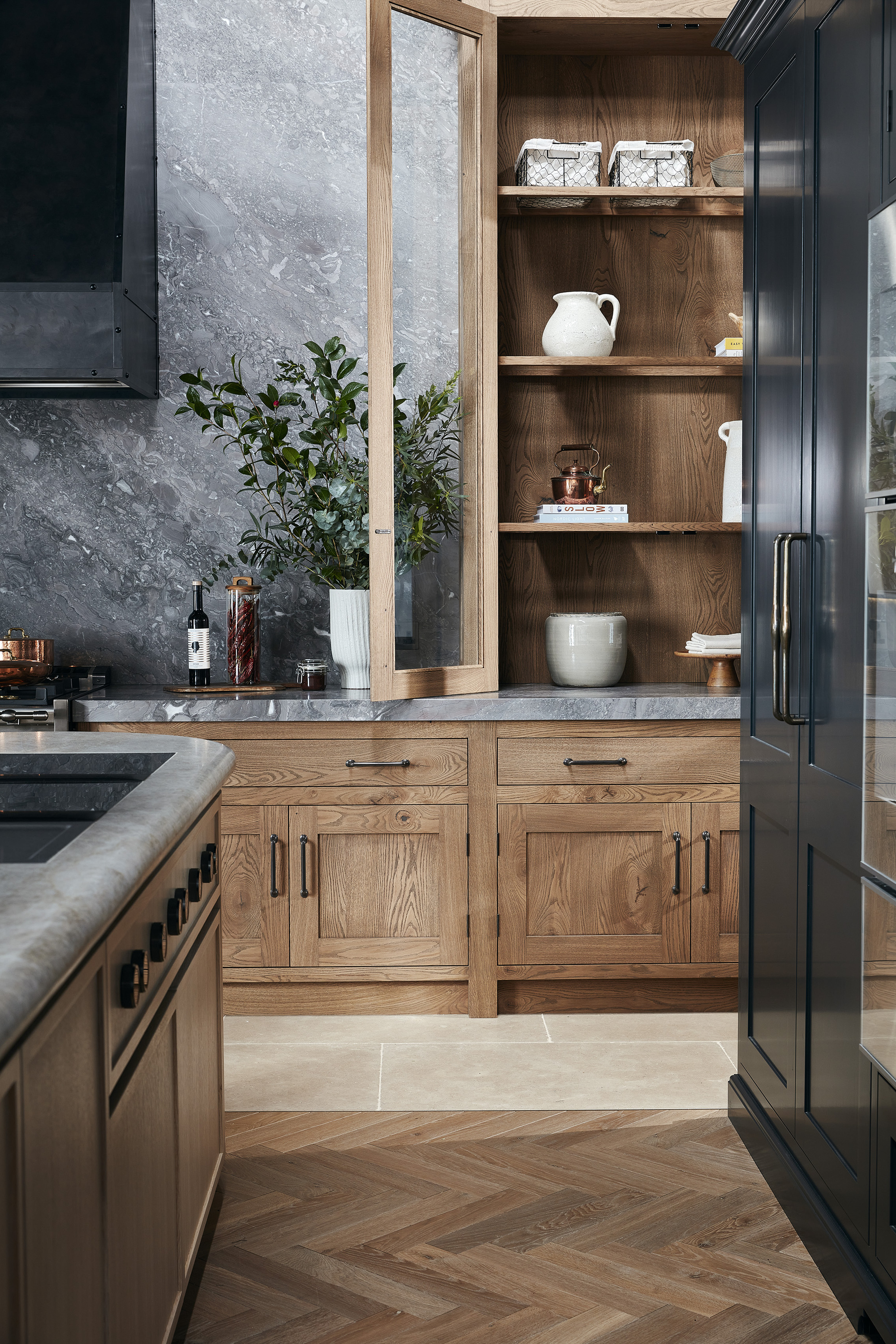 A modern kitchen with wooden cabinetry, grey countertops, and a mix of wooden herringbone flooring and large format tiles