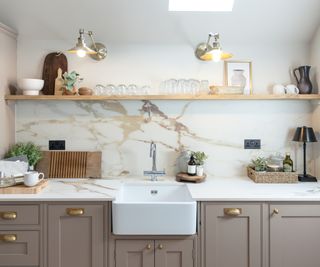 metal wall lights above wooden shelf and white butler sink in kitchen with mushroom coloured units and marble veined brown and white splashback and worktop