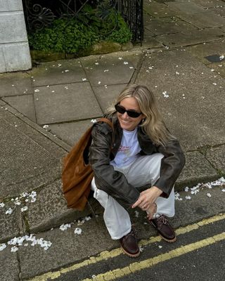 woman sitting on curb wearing brown leather jacket, white t-shirt, brown suede hobo bag, ecru pants, brown boat shoes, black sunglasses