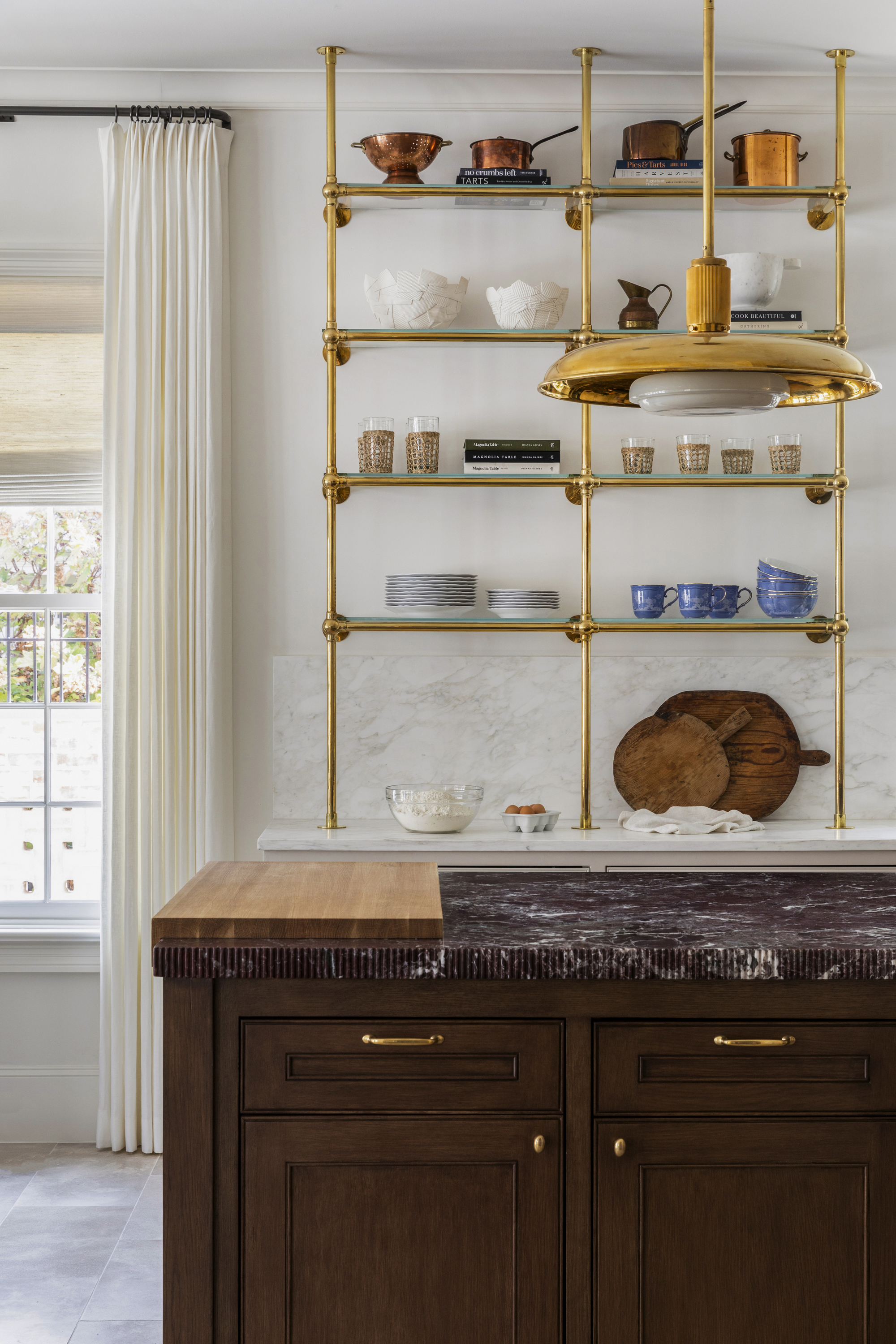 A kitchen with a wooden island with a red marble countertop and brass shelving on the wall storing kitchenware