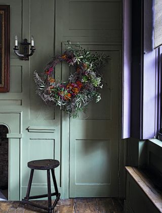 Wreath with a base of grey twigs and a large crescent effect of silvery green leaves, and bright yellow, orange, and purple blossoms, hanging over a door in the corner of a living room, next to the fireplace