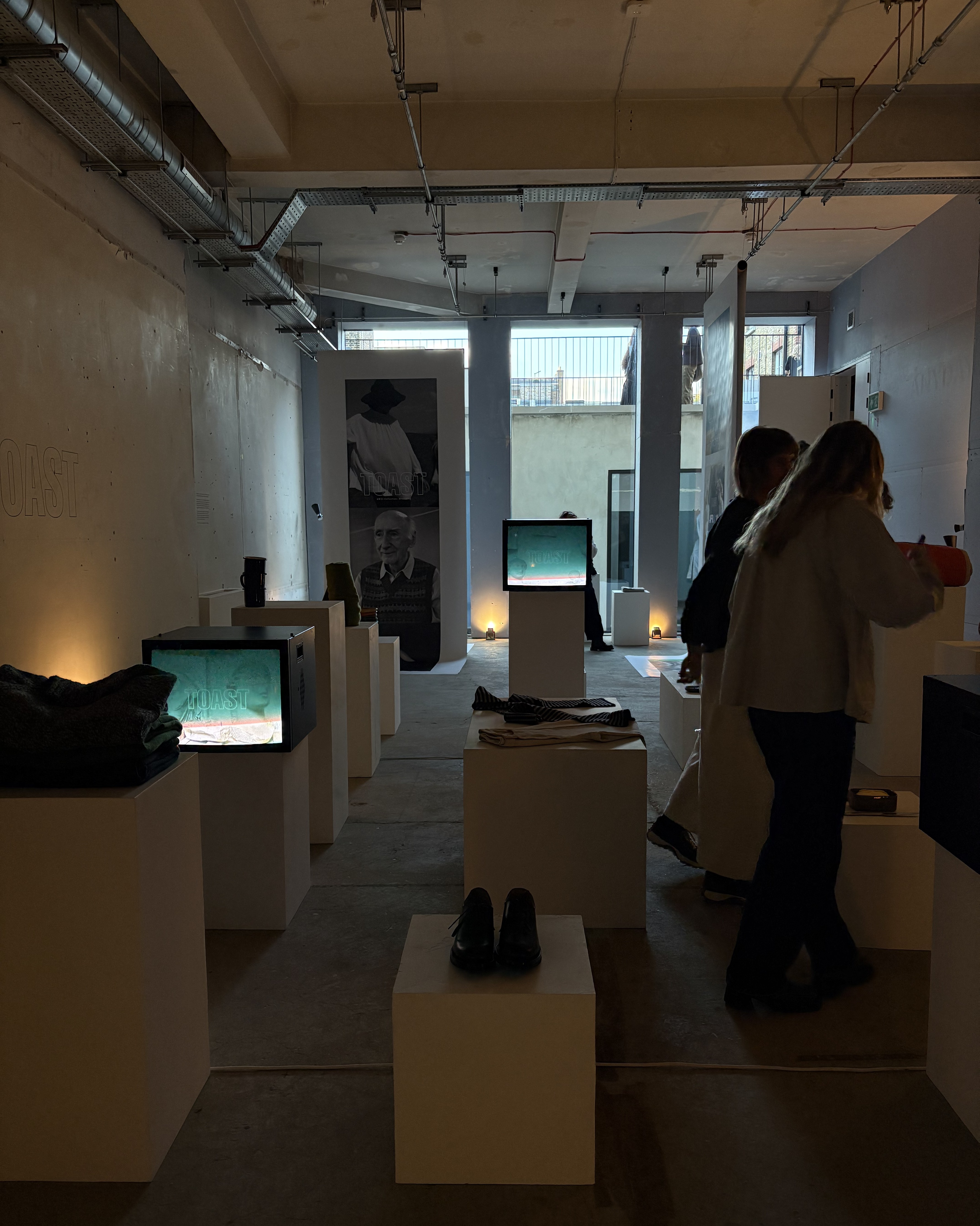 Installation view of a fashion and design exhibition featuring clothes, homeware, retro-looking tvs showcasing audiovisual works, large posters, and more.
