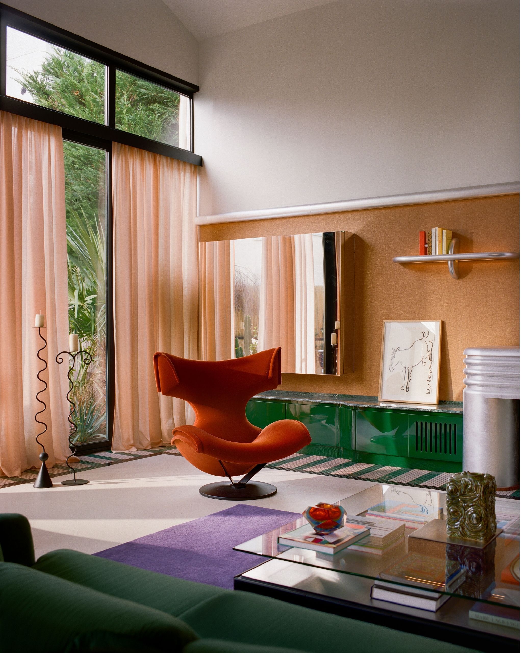 A living room with green cabinets, a chrome fireplace, a purple rug, a red accent chair, pink curtains, orange wallpaper, and a green sofa. 