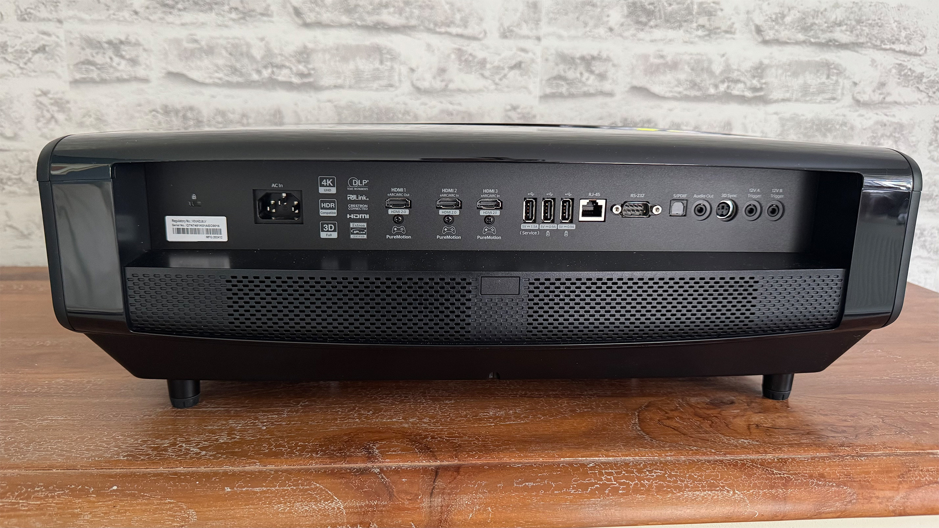 Optoma UHZ68LV home cinema projector on wooden surface in front of grey brick wall, rear of projector